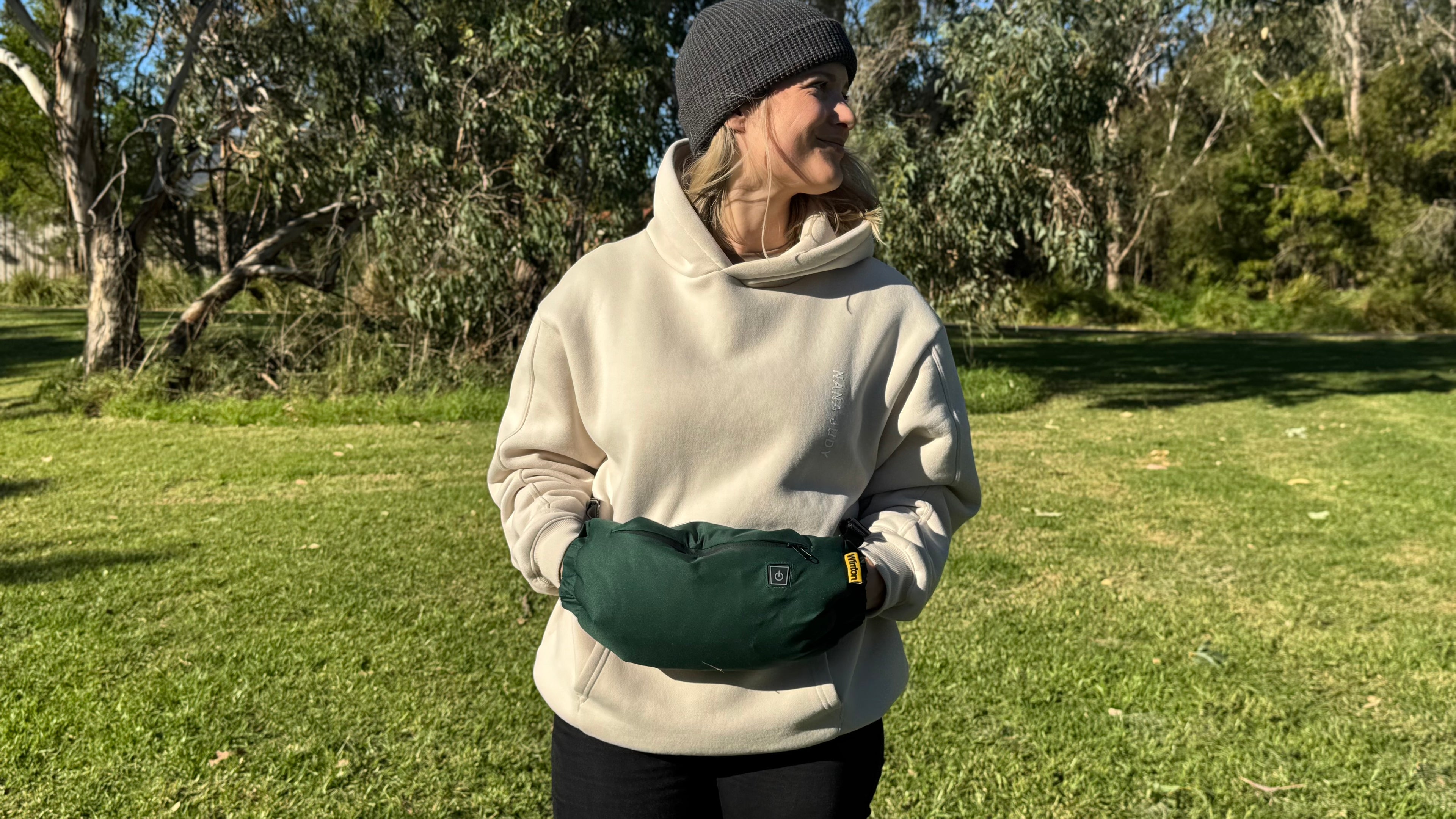 Person wearing a beige sweater and black pants with a green heated hand warmer in a grassy outdoor setting.