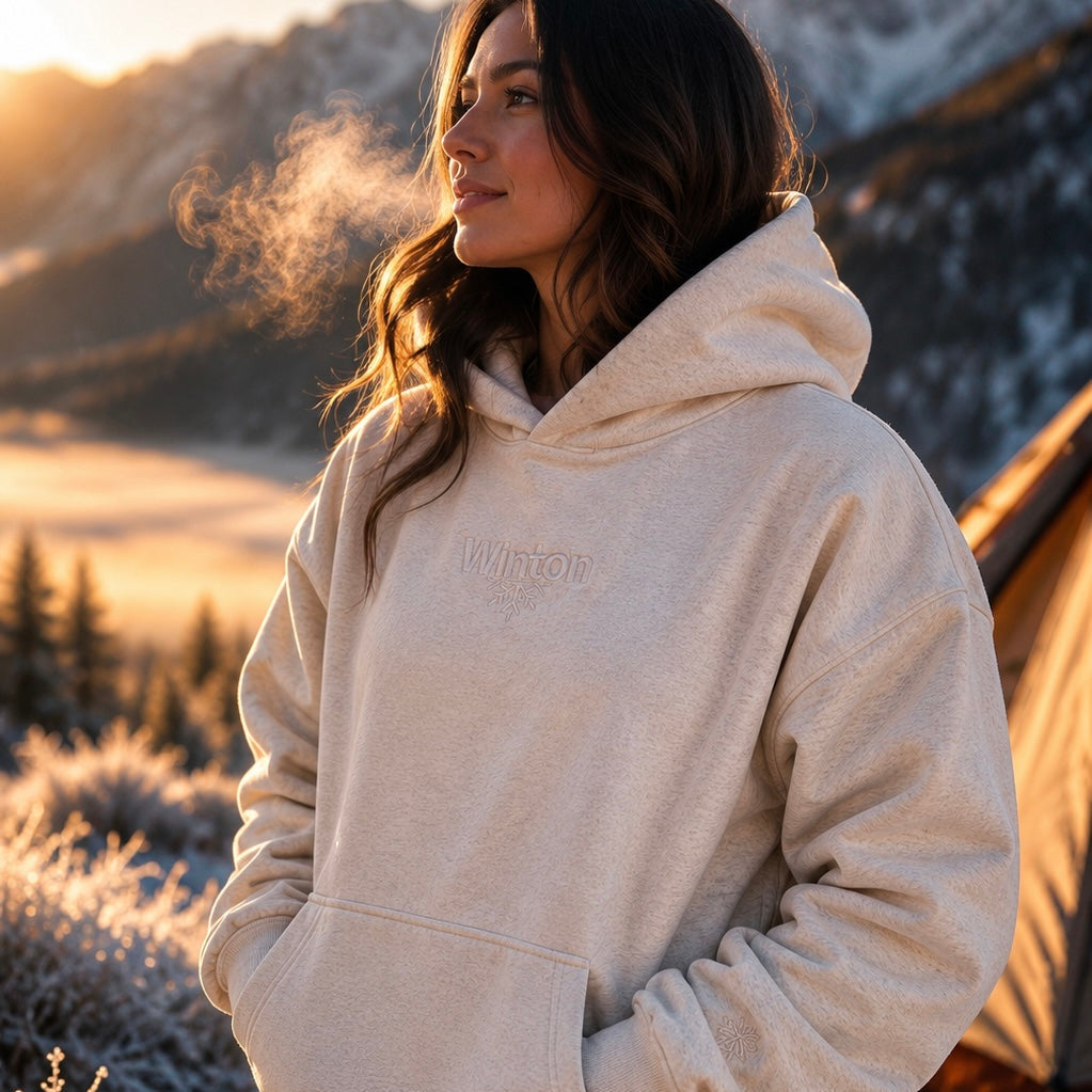 Woman wearing a beige hoodie standing in a snowy landscape with mountains in the background.